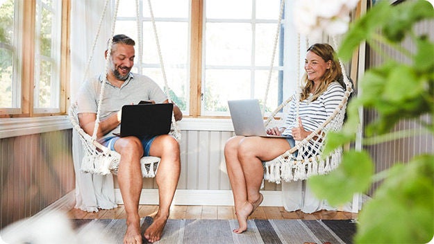 Two people are sitting in hanging chairs in a sunlit room with large windows. Both individuals are using laptops, suggesting they might be working or studying.