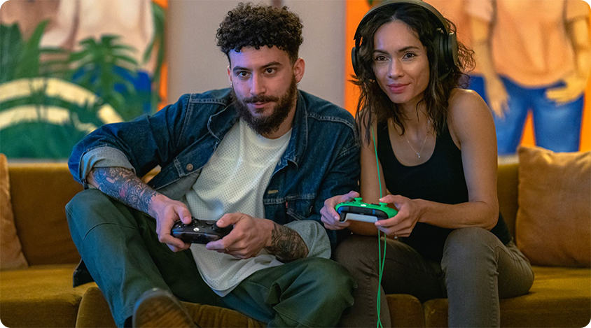 Two people sitting on a brown couch playing video games, each holding a controller. One person wears headphones and a black top, while the other wears a denim jacket over a white shirt. The background features colorful wall art with abstract figures and plants