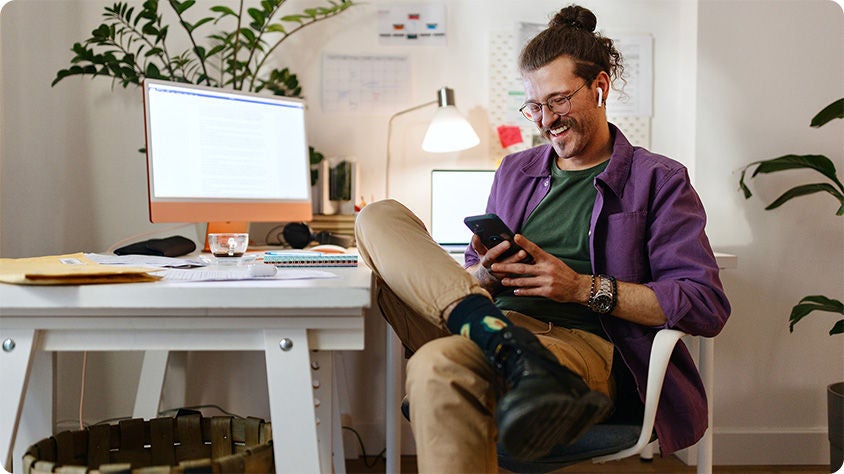 A smiling man with glasses and a bun hairstyle sits relaxed in a home office, looking at his phone while wearing wireless earbuds, with a desk, computer, and plants in the background.
