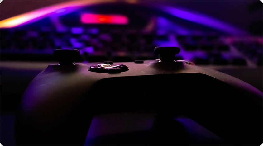 Close-up of a video game controller in front of a keyboard with purple and orange light 