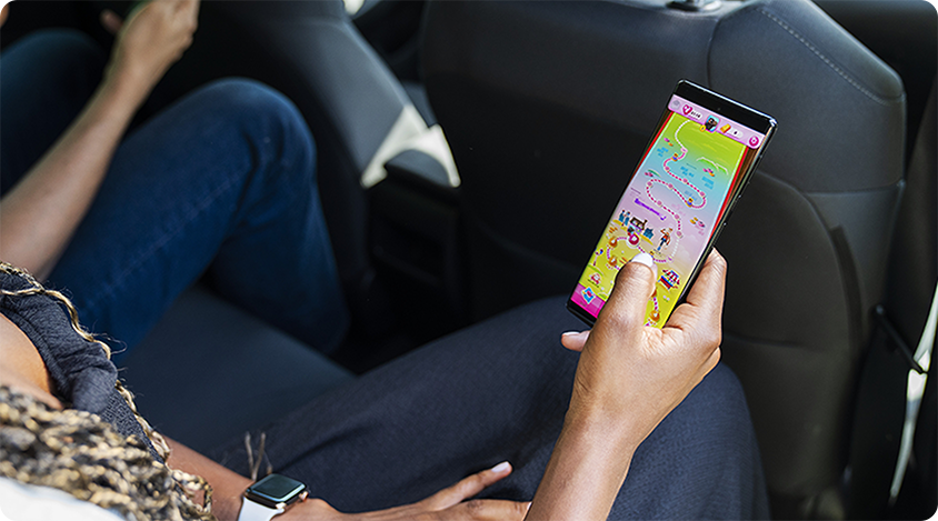 A person sits in the backseat of a car holding a smartphone that displays a colorful Candy Crush Saga map screen.