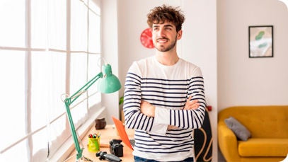 A person with crossed arms stands in front of a desk with various items and a green lamp, next to a window and a yellow sofa.