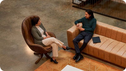 Two people in a modern office, one on a brown armchair with a laptop, and the other on an orange bench, facing each other.