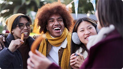 Four friends smile and gather as they enjoy churros at an outdoor holiday festival, dressed in winter clothing with festive decorations and palm trees in the background.