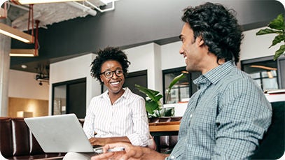 Two people sitting indoors. One person is holding a laptop and smiling.