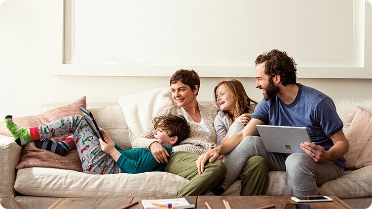 Two adults and two kids on a couch. One person is holding a laptop. One child is using a tablet.