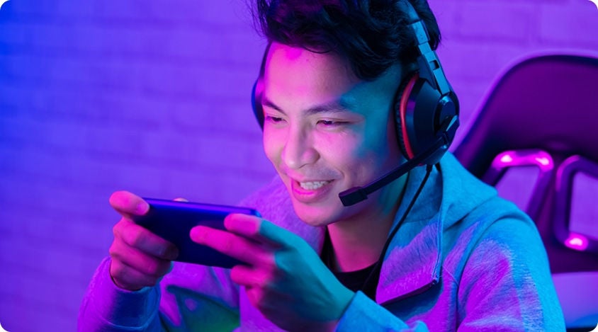 Young man wearing a gaming headset, smiling while playing a mobile game on his phone in a neon-lit room. 