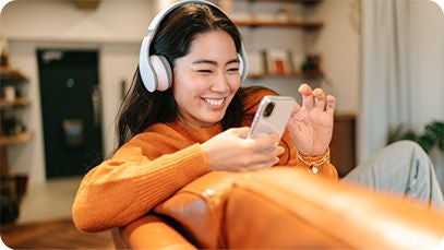 A person wearing headphones, sitting on an orange couch, and using a cellphone.