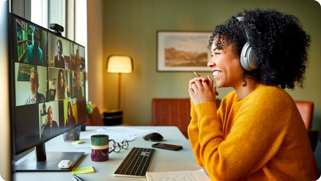 A person with curly hair and a yellow sweater sitting at a desk in front of a computer monitor having a video call with several people.