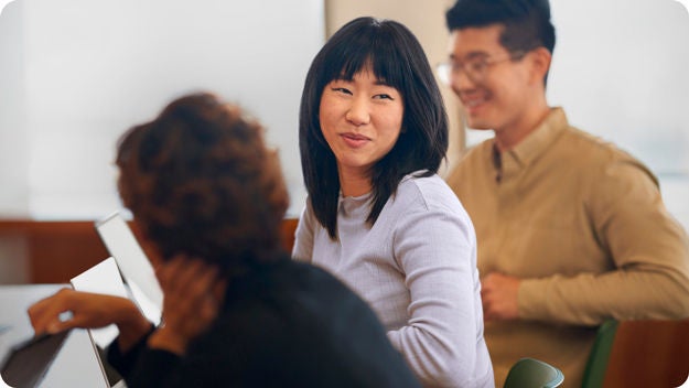 Three people in conversation at a table. One person is smiling and facing the camera, while the others engage nearby.