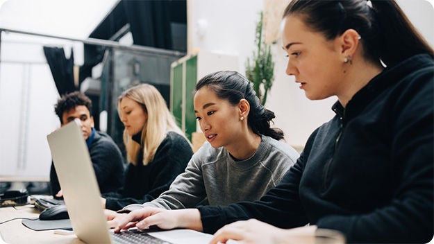 Four people sitting at a desk. The person in the front is using a laptop.