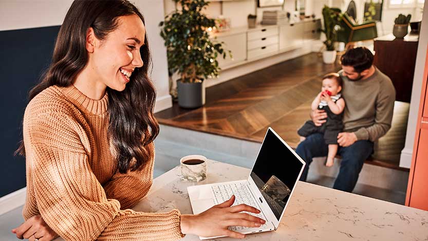 A woman smiles while working on a laptop computer while a man and a baby are interacting in the background