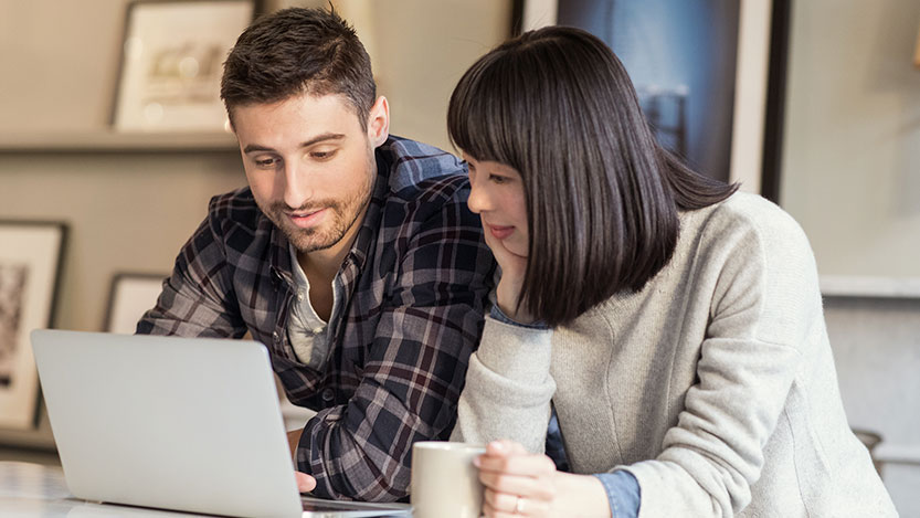 Two people work on a laptop computer while leaning against a table.