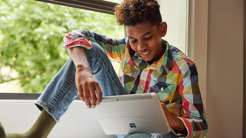 A young kid sits by a window while working on his tablet.