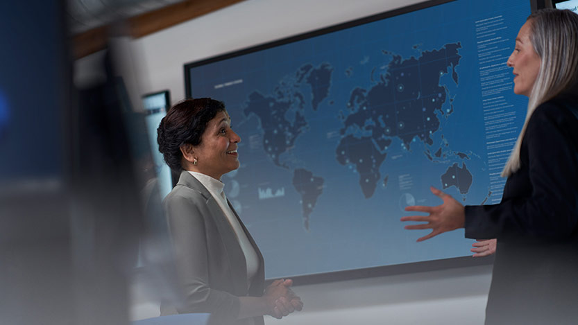 Two women facing each other talk in front of a world map in an office.