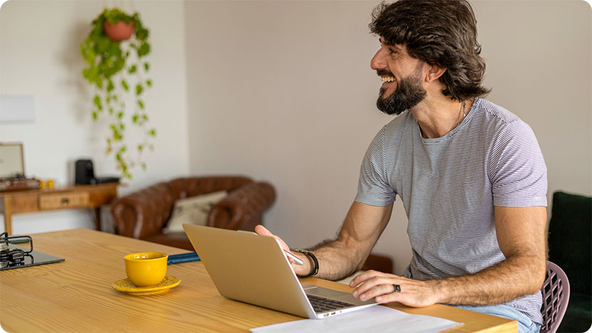 Person, die mit einem Laptop an einem Tisch sitzt, ein Smartphone in der Hand hält und lächelt.