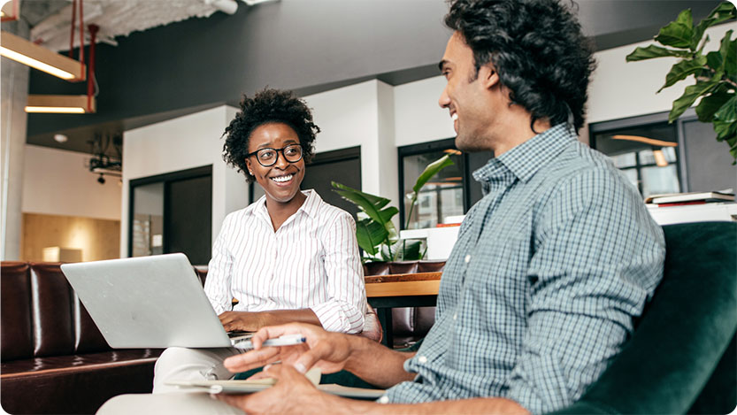 Two people sitting in a room smiling at each other. One person is holding a laptop.