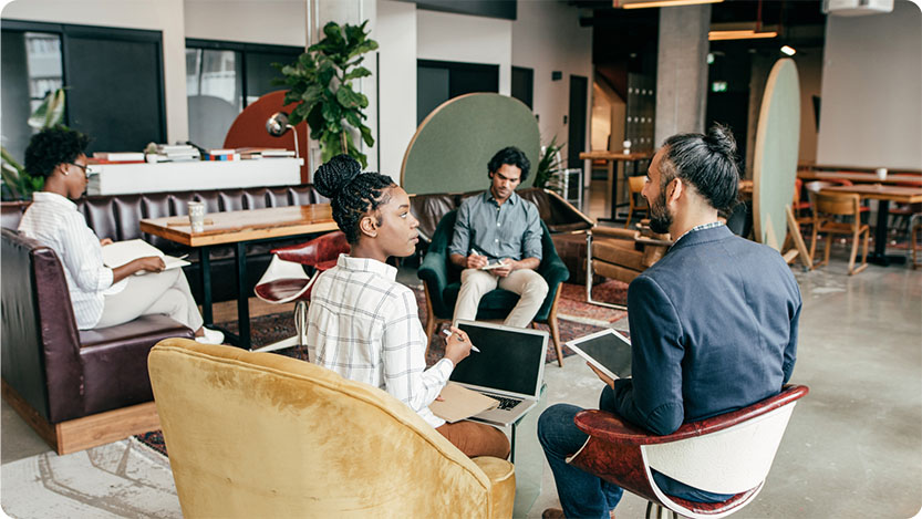 A group of people in a modern office space engaged in a discussion. Two are seated on mustard yellow armchairs facing each other with laptops, while another person sits on a green sofa with a laptop. In the background, another person is seated at a table working independently.