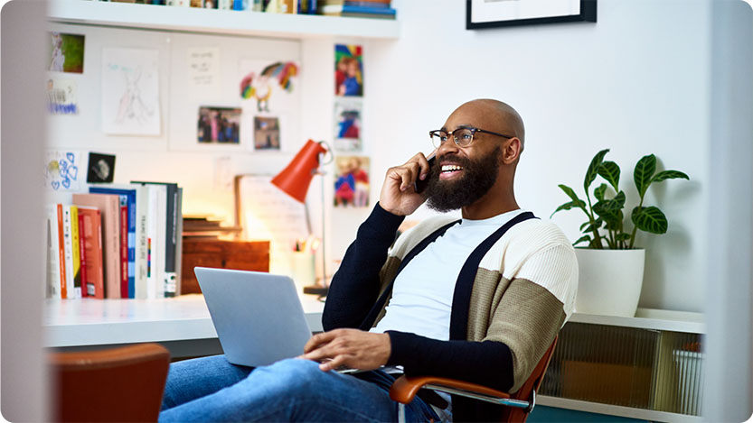 A person is sitting in a modern, well-lit office space, holding a phone to their ear and using a laptop.