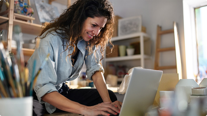 A person with long, curly hair is leaning over a table in a well-lit room filled with various art supplies and shelves. The individual appears to be focused on their work, which involves using a laptop and other materials on the table