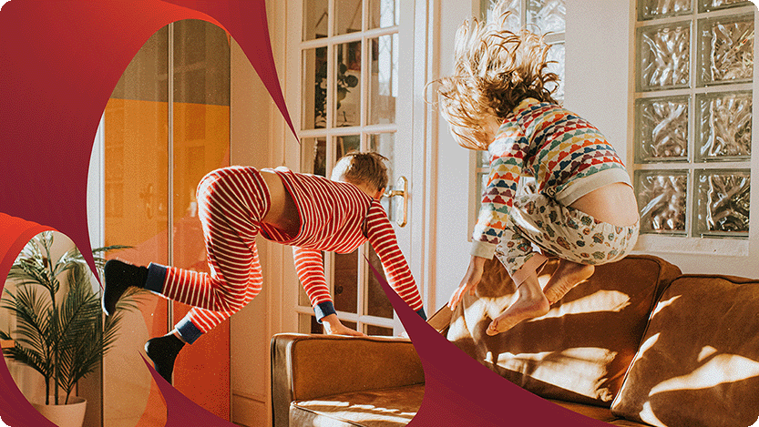 Two children are captured mid-air while jumping on a brown couch in a brightly lit room. One child is wearing red and white striped pajamas, while the other is dressed in colorful patterned pajamas.