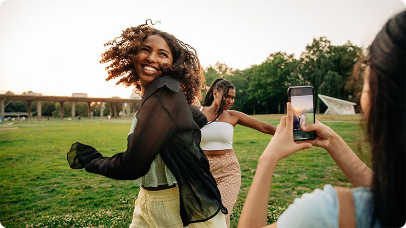 Three people in a park at sunset; one person takes a photo with a smartphone while two others dance or spin on the grass, with trees and a bridge in the background.