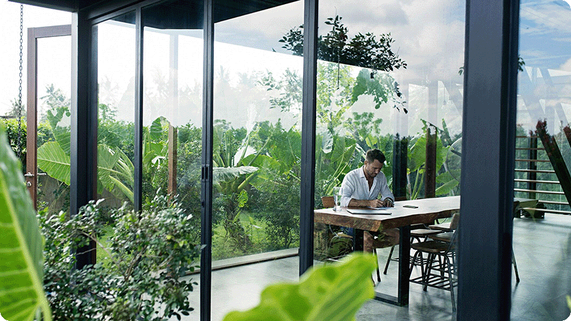 A mature man is sitting at a table in front of a lush garden, writing.