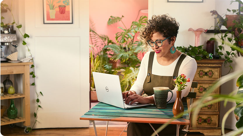 A person typing on a laptop at a small table with a mug and a vase of flowers, surrounded by plants and framed artwork in a cozy indoor setting.