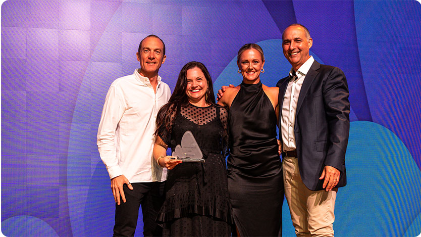 Group photo of four individuals in front of a blue and purple backdrop, with one person holding an award; capturing a celebratory moment.