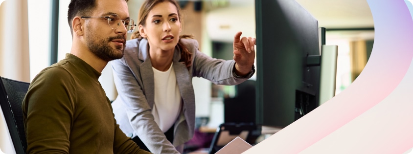 Two colleagues collaborate at a workstation in a modern office, with one seated at a desk typing on a computer and another standing beside them, pointing at the monitor as they review work together.