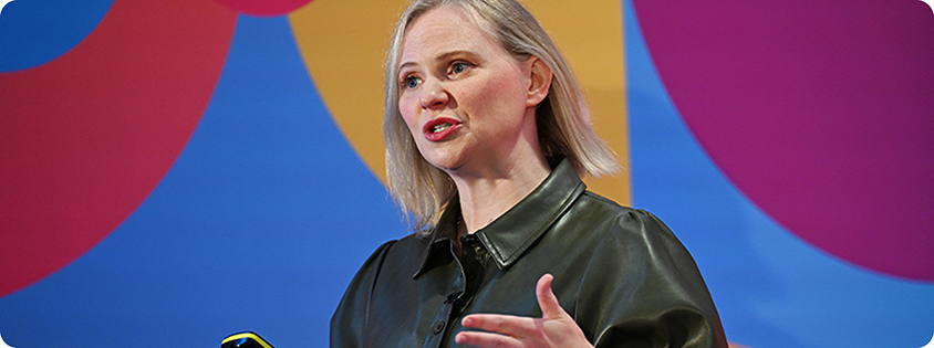 Melinda Spence presenting onstage at Advertising Week Europe 2024 with a phone in hand in front of a colorful backdrop..