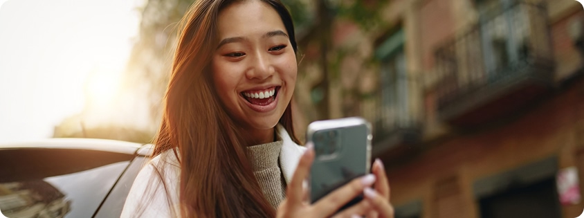 Woman standing outdoors and smiling at her smartphone in a sunlit urban setting 