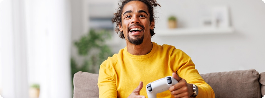 Man sitting on his couch smiling and tilting his body along with the controller he is holding as he plays a video game. 