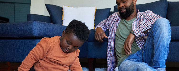 A father sits on the floor and leans on the couch while watching his son play.