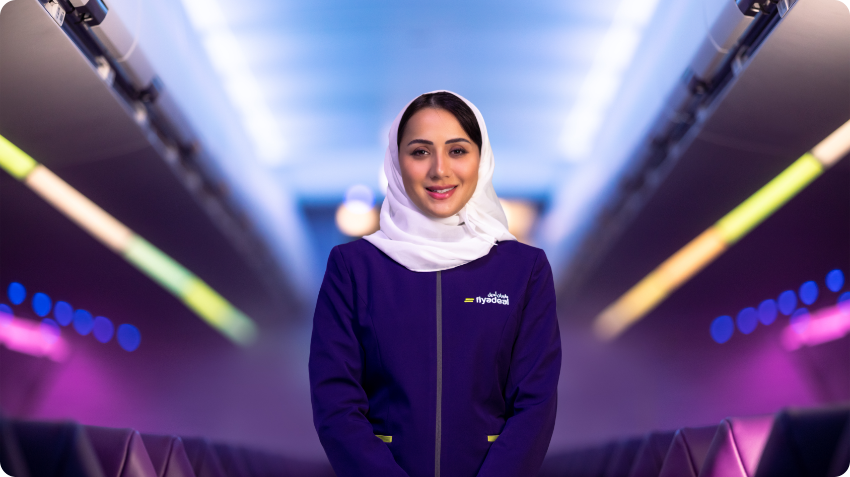 A person wearing a purple uniform with a white headscarf stands in the aisle of an airplane. The uniform features the 'flyadeal' logo. The airplane cabin has seats on both sides and overhead compartments, illuminated with colorful lighting in blue, green, and pink hues.