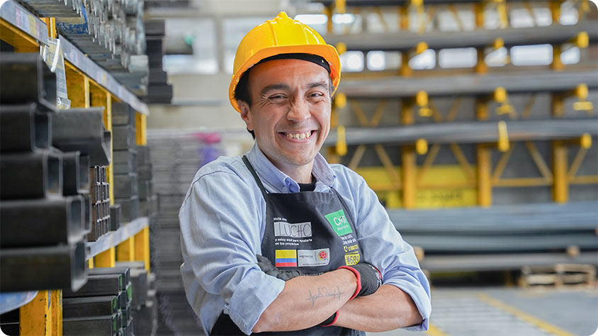 A person in a yellow hard hat and light blue shirt stands with arms crossed in an industrial warehouse. They wear a black apron displaying badges, including 'Hola Soy Lucho' and '#689'. Their face is blurred. Behind them are shelves stocked with metal beams and industrial supplies.