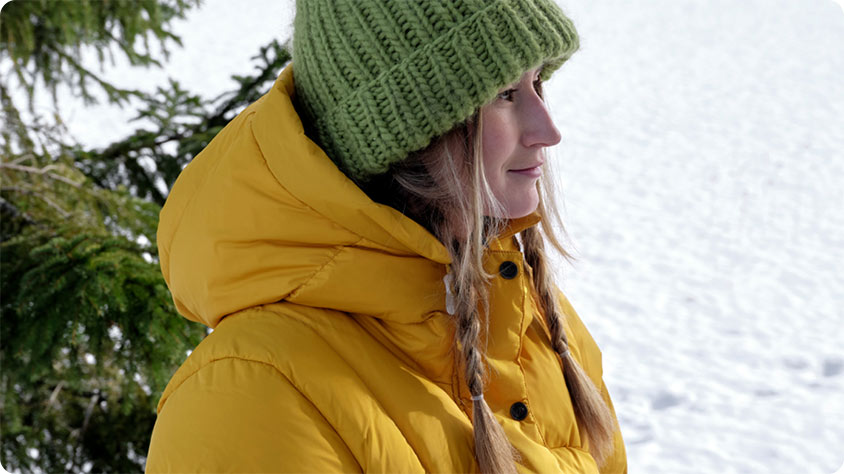 A person wearing a bright yellow puffer jacket and a green knitted beanie stands outdoors in a snowy environment.