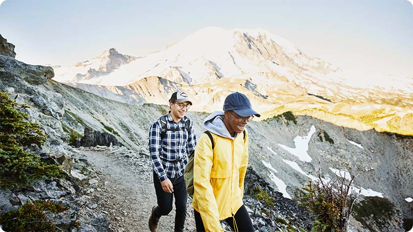 Two people hiking on a mountain trail with a large, snow-capped mountain in the background under a clear blue sky.