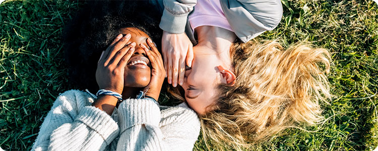 Two people laying on the grass. One is whispering in the other one’s ear.