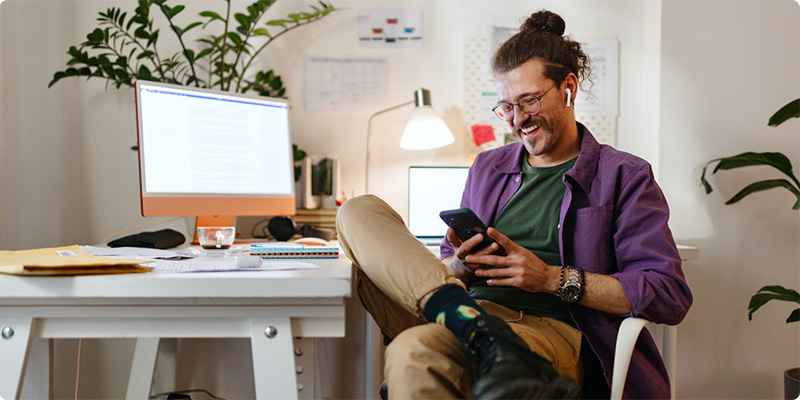 A man is smiling while looking down at his phone at his home office.